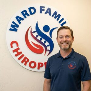 Dr. Jason Ward smiling in a navy polo shirt, standing proudly in front of the large circular Ward Family Chiropractic sign on the office wall.